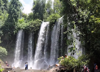 The wide, cascading upper falls of Phnom Kulen surrounded by green trees and volcanic rocks in Cambodia.
